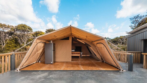 Een grote, open tent met een houten binnenkant, een klein bankje en een koelkast.