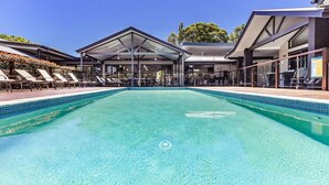 Una piscina con sillas de descanso y una zona cubierta, rodeada de edificios con grandes ventanales y un cielo despejado.