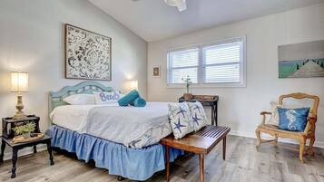 A bedroom with a bed, a chair, a table, a lamp, and artwork on the walls.