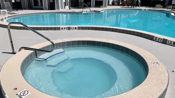 A hot tub with a pool in the background, surrounded by lounge chairs and umbrellas.