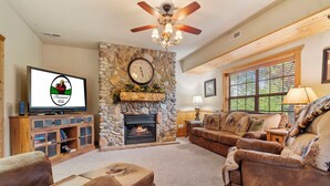 A living room with a stone fireplace, a television, and a ceiling fan. - Black Bear Lodge: Lovely 2BR+Loft Cedar Cabin Near Shows/Eateries/Ticket & Golf DEALS! (Branson)