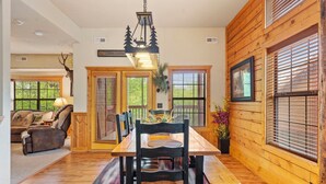 A dining area with a wooden table and chairs, a chandelier, and a view of the outdoors through large windows. - Black Bear Lodge: Lovely 2BR+Loft Cedar Cabin Near Shows/Eateries/Ticket & Golf DEALS! (Branson)