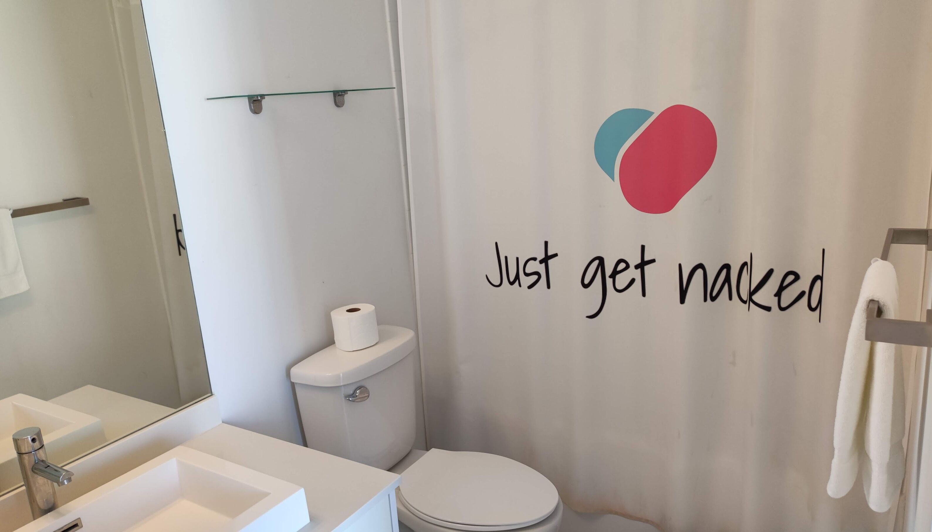 Et badeværelse med toilet, håndvask og bruseforhæng med hjerte-design samt teksten "Just get naked".