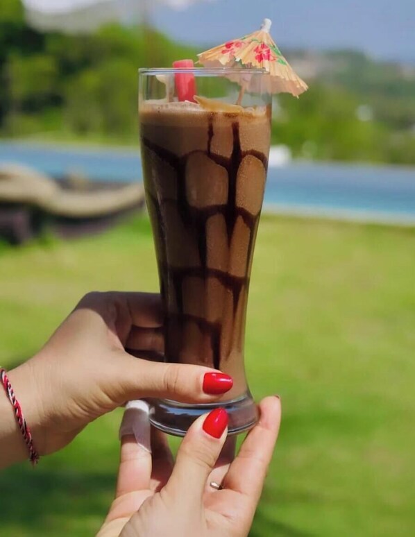 A tall glass of iced coffee with a straw and a small umbrella on top, held by hands with red nail polish. -  Uluwatu Sunset Hills (Pecatu)