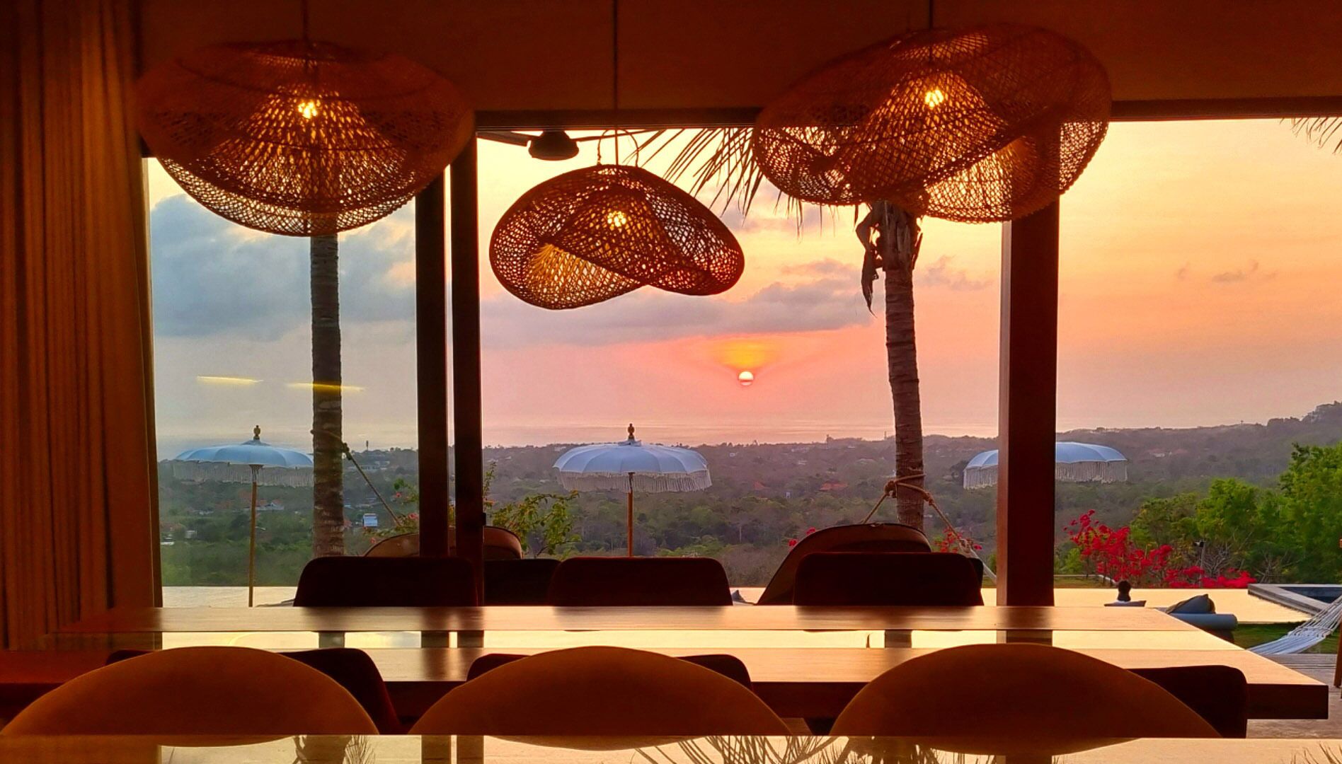 A restaurant with a view of the sunset over a cityscape, featuring large woven umbrellas and a table with chairs.