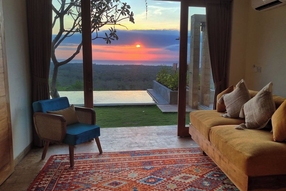 Una habitación con vista a un atardecer sobre una masa de agua, un sillón azul, un sofá amarillo y una alfombra estampada.