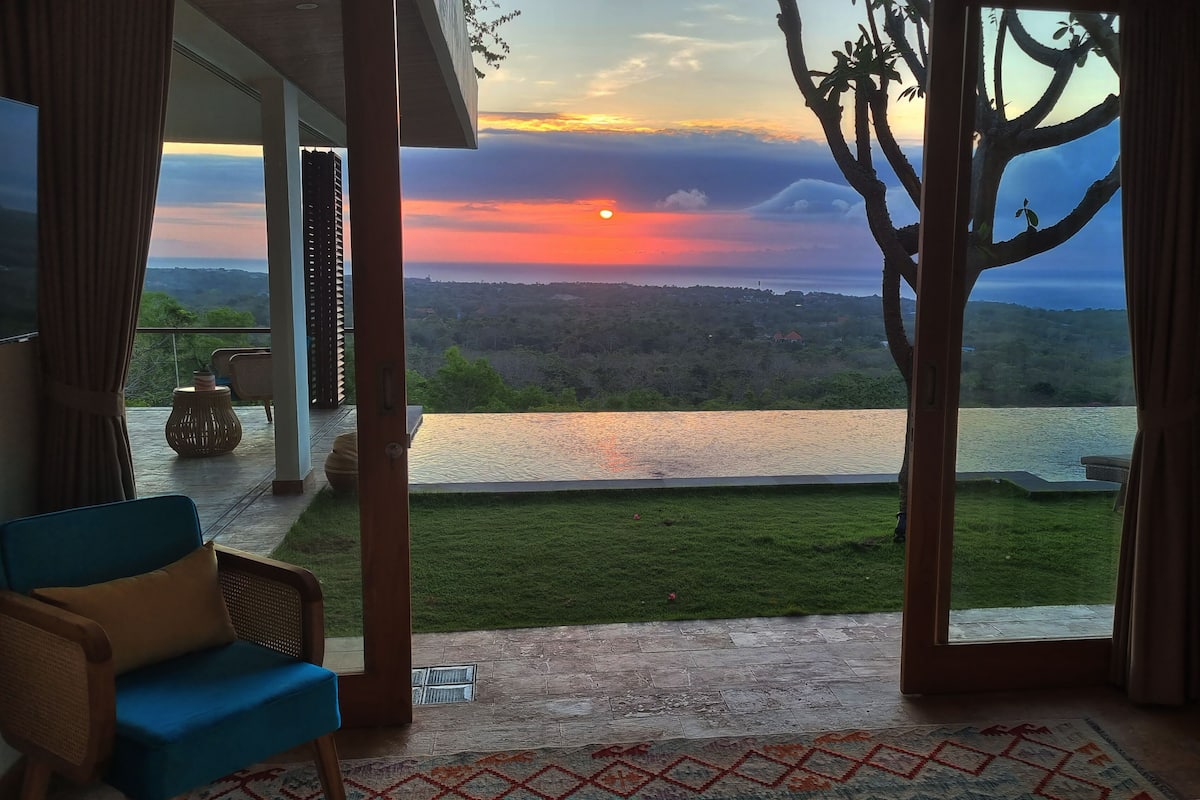 Una habitación con vistas a un atardecer sobre el agua, una silla azul y una alfombra estampada.