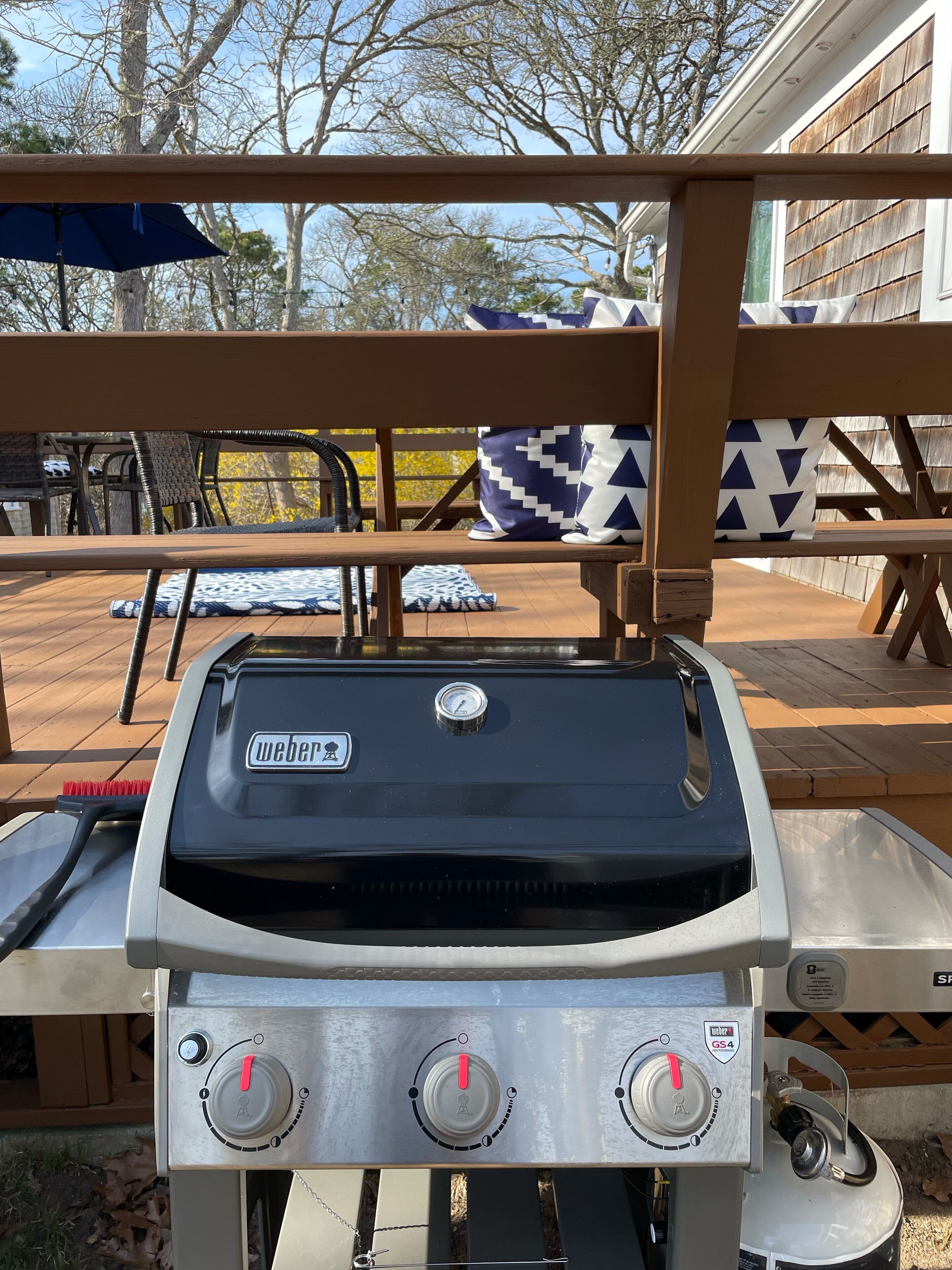 En Weber gasgrill med digitalt termometer og kontrolknapper, placeret på en udendørs terrasse med træmøbler og en blå parasol.