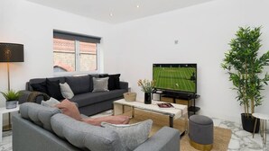 Ein modernes Wohnzimmer mit einem grauen Sofa, einem Couchtisch, einem Fernseher, der ein Sportspiel zeigt, und einer großen Küstpflanze.