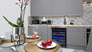 Eine moderne Küche mit grauen Schränken, einer Mikrowelle, einer Kaffeemaschine und einer Ananas auf der Arbeitsplatte. Es gibt einen Glastisch mit einer gewebten Tischmatte, eine Glasvase mit Orchideen und einen Teller mit Äpfeln.