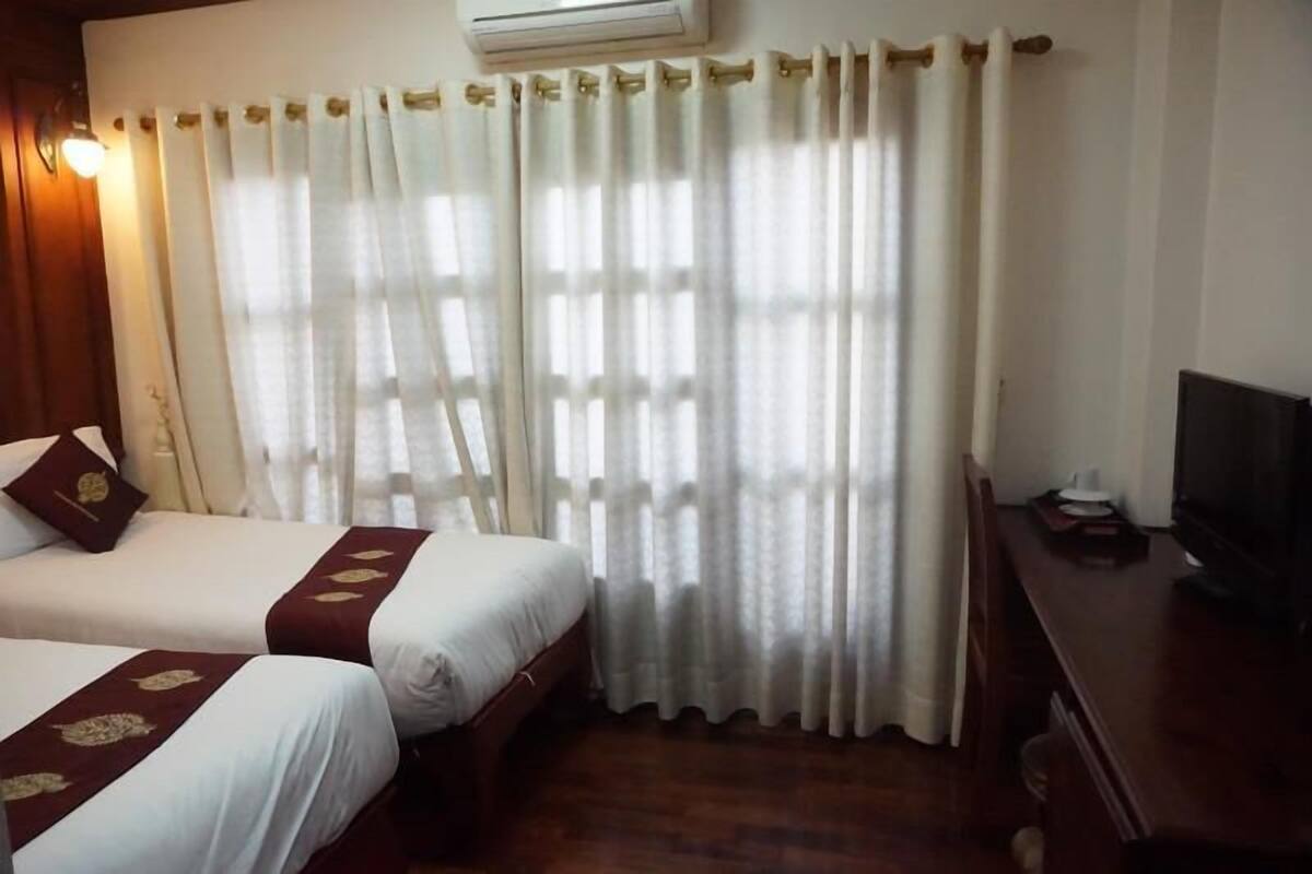 Une chambre d’hôtel avec deux lits, un téléviseur posé sur une table en bois et une fenêtre ornée de rideaux légers.