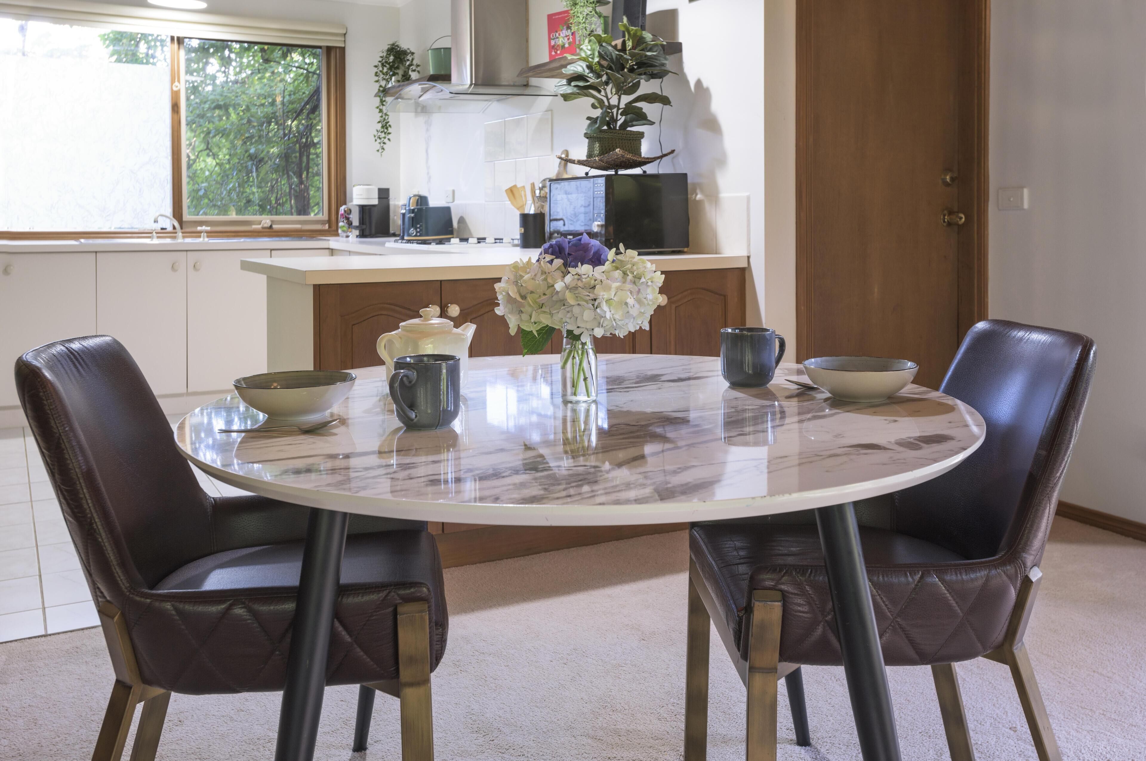 Un área de comedor con una mesa redonda, dos sillas, un jarrón con flores y electrodomésticos de cocina al fondo.