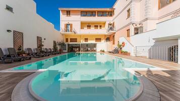A swimming pool with lounge chairs and a covered seating area, surrounded by a building with balconies and a white exterior.