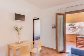 Ein Zimmer mit Schreibtisch aus Holz, zwei Stühlen, einem an der Wand befestigten Fernseher, einem großen Fenster mit Blick auf Gebäude und einer Glastür, die zu einem Außenbereich führt.