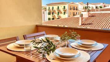 A wooden table set with plates, glasses, and olive branches on a balcony with a view of terracotta-roofed buildings.