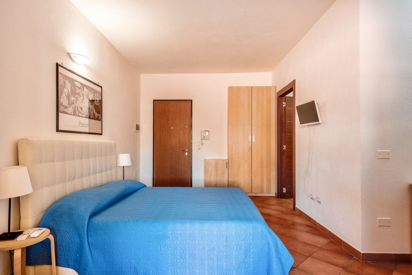 Een hotelkamer met een bed, nachtkastje met lamp, een lijst met afbeelding aan de muur, een garderobe en een televisie.