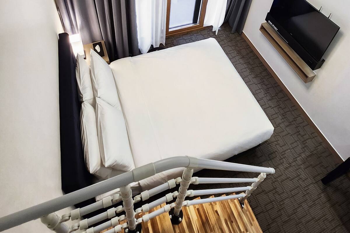 Ein modernes Hotelzimmer mit Bett, Fernseher, Holzboden und einem Fenster mit Vorhängen.