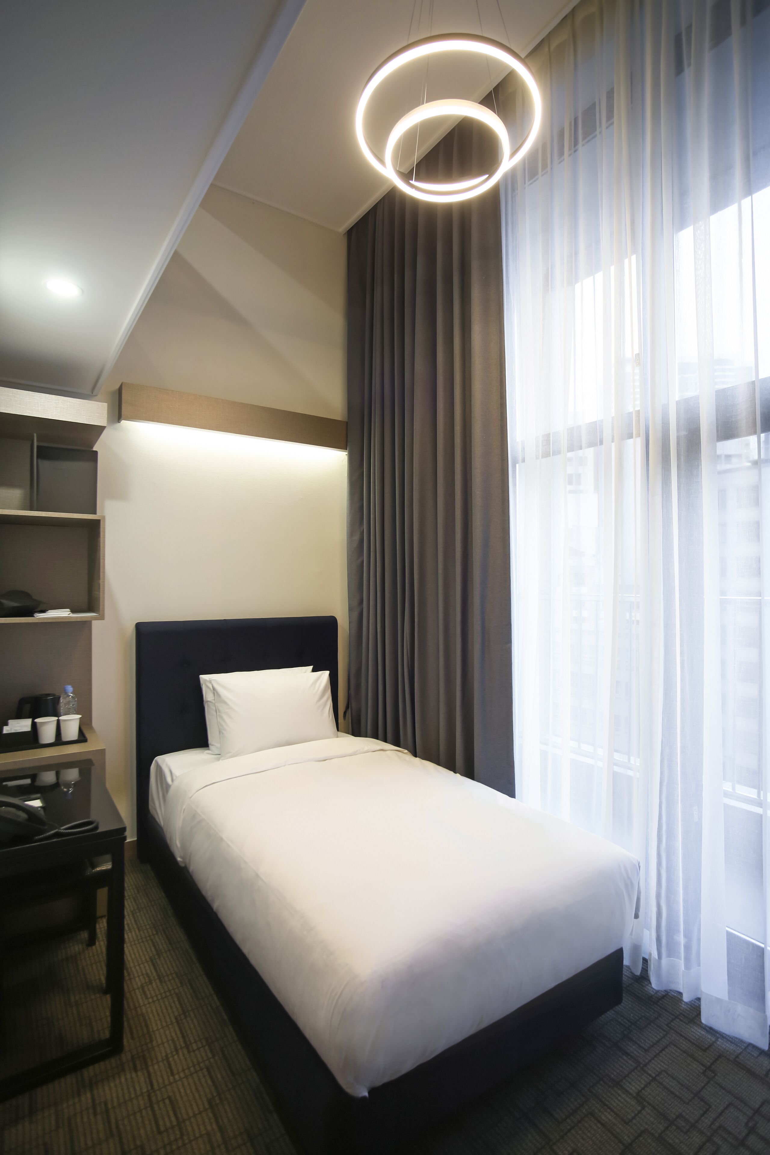 A hotel room with a large bed, a desk with a lamp, and a view of the city through sheer curtains.