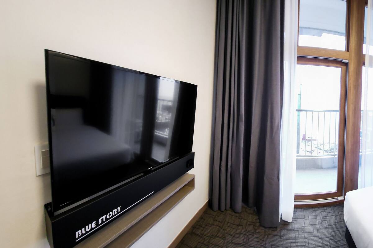 Ein Hotelzimmer mit Flachbildfernseher, einem Bett mit weißer Bettwäsche, einem Nachttisch mit Lampe und Blick ins Freie durch ein großes Fenster mit Vorhängen.