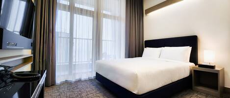 Habitación de hotel con una cama grande, un televisor en un soporte, un escritorio con lámpara y vistas al exterior a través de cortinas translúcidas.