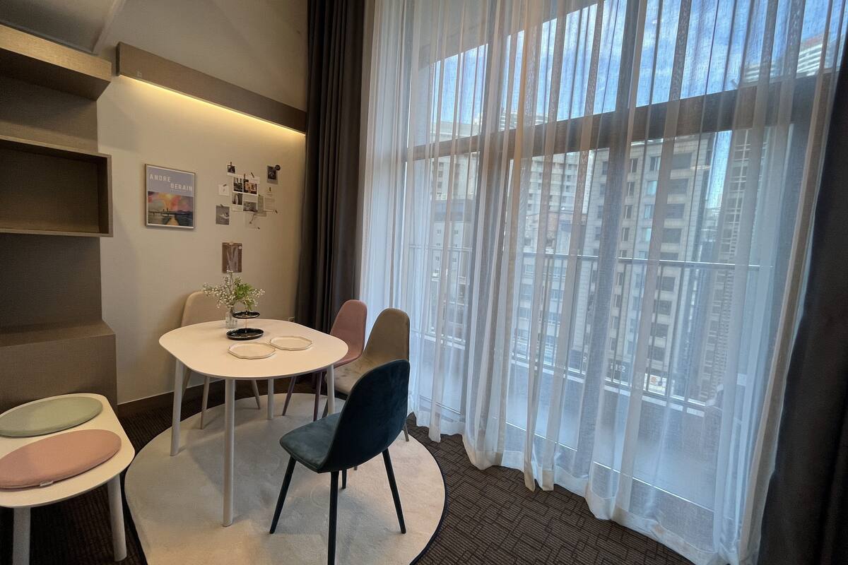 Ein modernes Hotelzimmer mit einem runden Esstisch, zwei Stühlen, einem kleinen Beistelltisch und einem großen Fenster mit durchsichtigen Vorhängen.