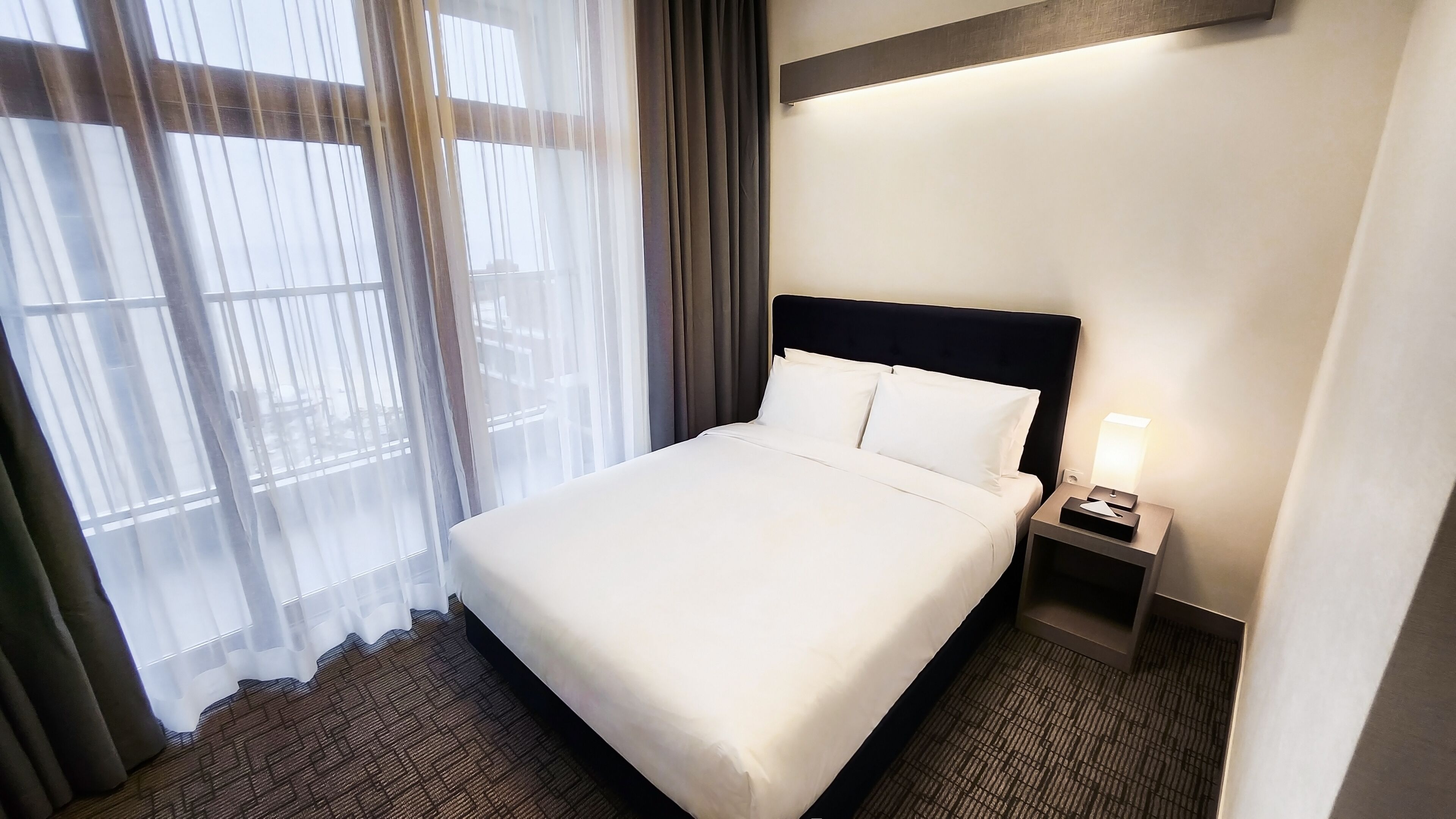 A hotel room with a large bed, a nightstand with a lamp, sheer curtains, and a view through the window.
