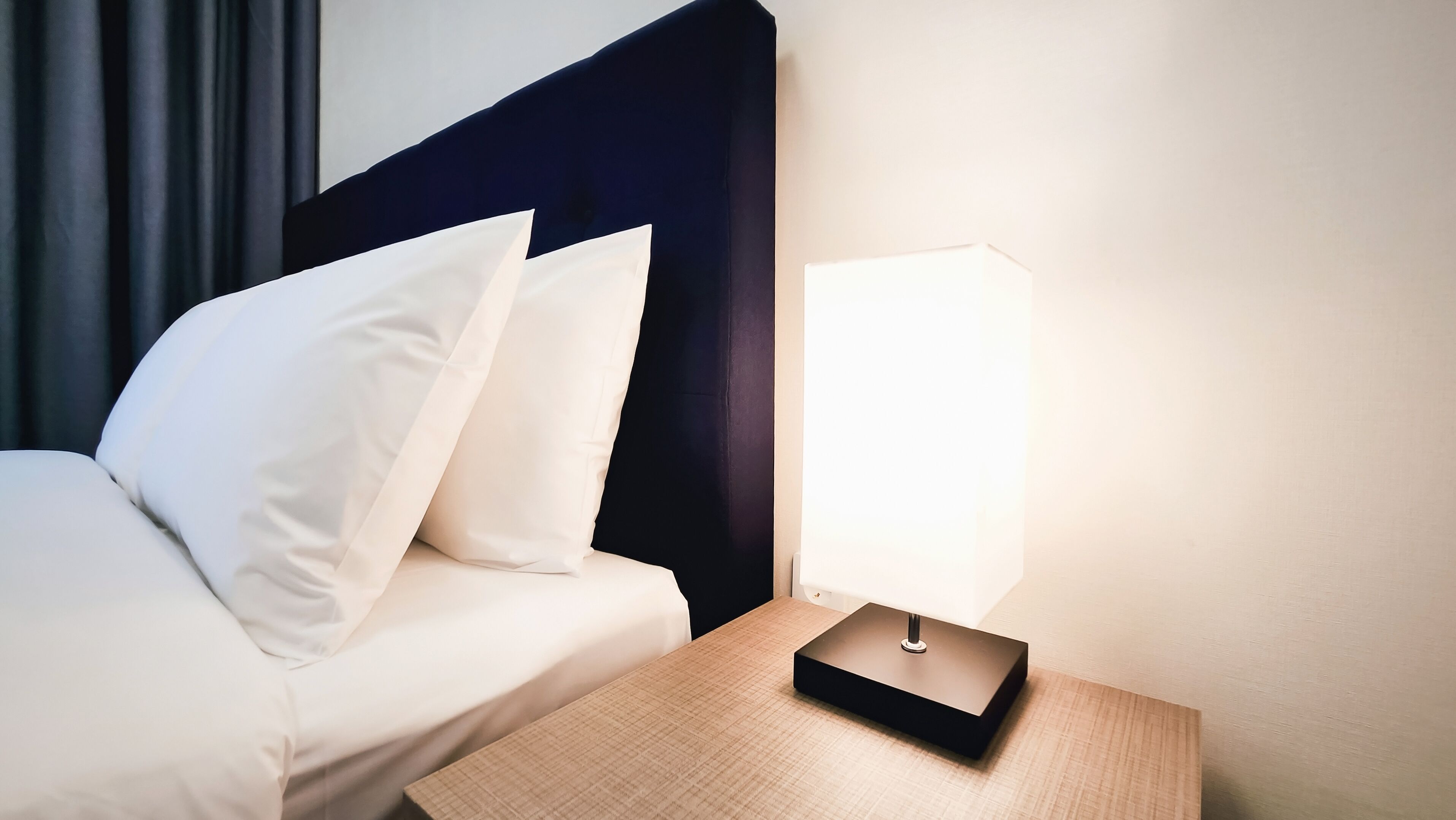 En pænt redt seng med hvide sengetæppe og puder, et natbord med lampe, og en mørk sengegavl.