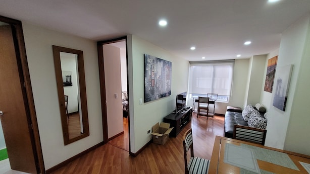 Amplia sala de estar con piso de madera, una mesa de comedor, un sofá y un televisor. Hay un ventanal con persianas, una puerta que conduce a otra habitación y un cuadro en la pared.