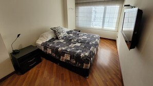 Un dormitorio con cama, mesita de noche, lámpara, ventana con cortinas y televisor montado en la pared.