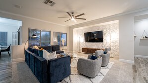 A modern living room with a dark blue sectional sofa, grey armchairs, a flat-screen TV mounted on a textured wall, and a ceiling fan.