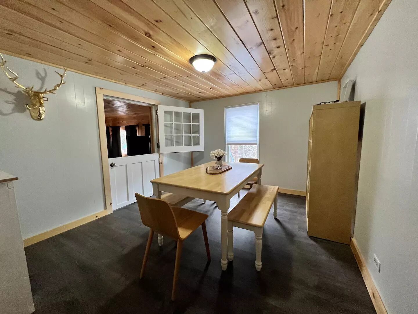 A room with a wooden ceiling, a dining table with chairs, a bench, a refrigerator, and a door leading to another room.