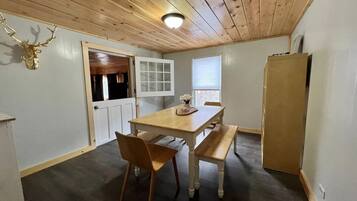 A room with a wooden ceiling, a dining table with chairs, a bench, a refrigerator, and a door leading to another room.