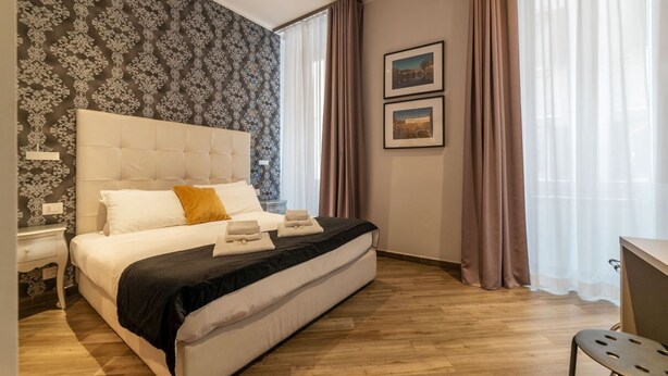 A bedroom with a large bed, a nightstand, a desk, and a window with curtains. - Frattina 2 - FromHometoRome (Roma)