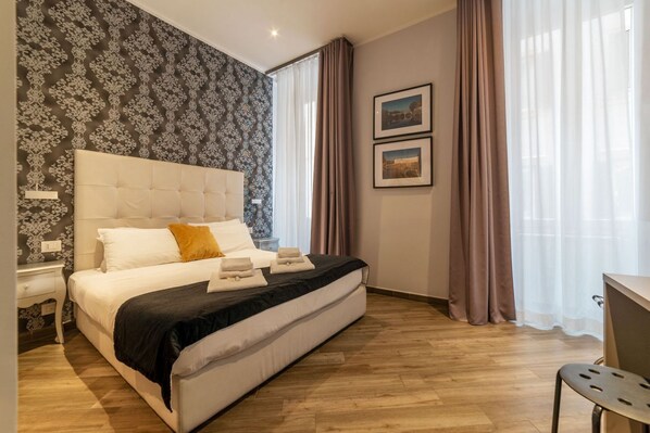 A bedroom with a large bed, a nightstand, a desk, and a window with curtains.