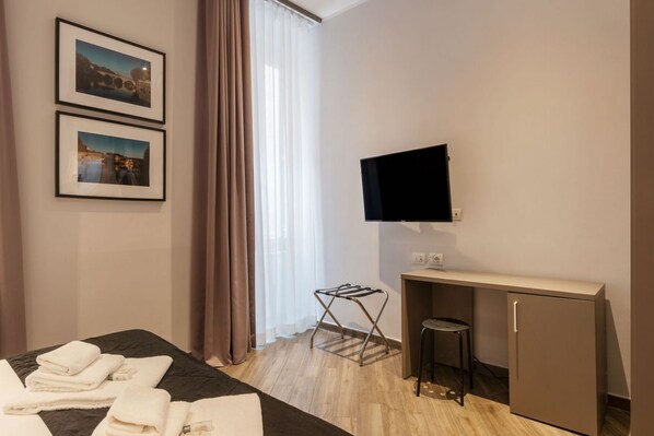 A hotel room with a bed, a desk with a TV, a small stool, and a drying rack.