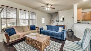 A spacious living room with a beige sofa, a blue sofa, a wooden coffee table, and a striped armchair. A ceiling fan is visible, and there is a staircase leading to an upper level. - Spacious Farmhouse  Townhome*Walk to AT&T Stadium* (Arlington)