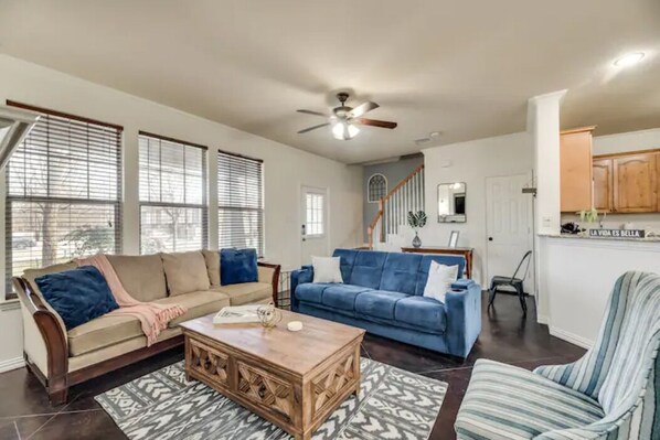 Ein geräumiges Wohnzimmer mit einem beige-farbenen Sofa, einem blauen Sofa, einem Holztisch und einem gestreiften Sessel. Ein Deckenventilator ist sichtbar, und es führt eine Treppe in eine obere Etage.