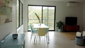 Ampla sala de estar com mesa de jantar, cadeiras, sofá, TV e uma grande janela com vista para árvores.