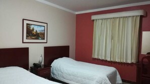 Habitación de hotel con dos camas individuales, una mesita de noche, una lámpara, un cuadro en la pared y una ventana con cortinas.