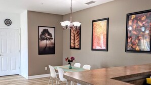 A modern kitchen with a wooden countertop, white chairs, and a dining table. There are framed artworks on the walls and a chandelier above the table.