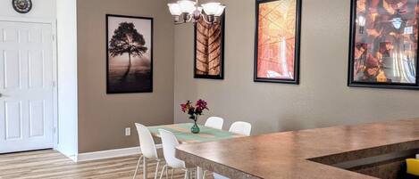 A modern kitchen with a wooden countertop, white chairs, and a dining table. There are framed artworks on the walls and a chandelier above the table.