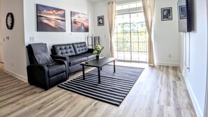 A modern living room with a black leather sofa, a coffee table, and a striped rug. There are two large windows with curtains, a ceiling fan, and a flat-screen TV mounted on the wall.