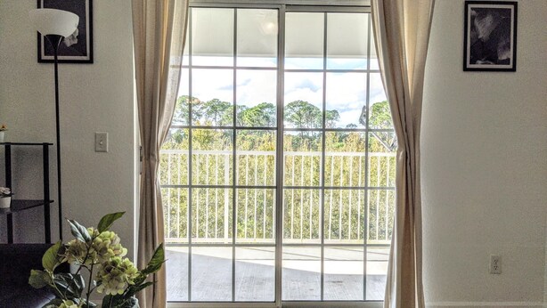 A room with a large sliding glass door leading to a balcony with a metal railing. There is a potted plant on the floor and a small table with a lamp.