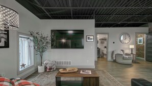 A modern living room with a large flat-screen TV, a fireplace, a comfortable sofa, and a dining area with a wooden table. - The 418 ~ An 1866 Build with Modern Comforts (Fort Wayne)