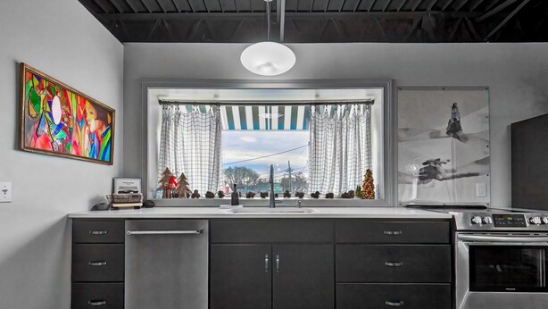 A modern kitchen with a built-in dishwasher, stainless steel appliances, a large window with curtains, and a colorful abstract painting on the wall. - The 418 ~ An 1866 Build with Modern Comforts (Fort Wayne)