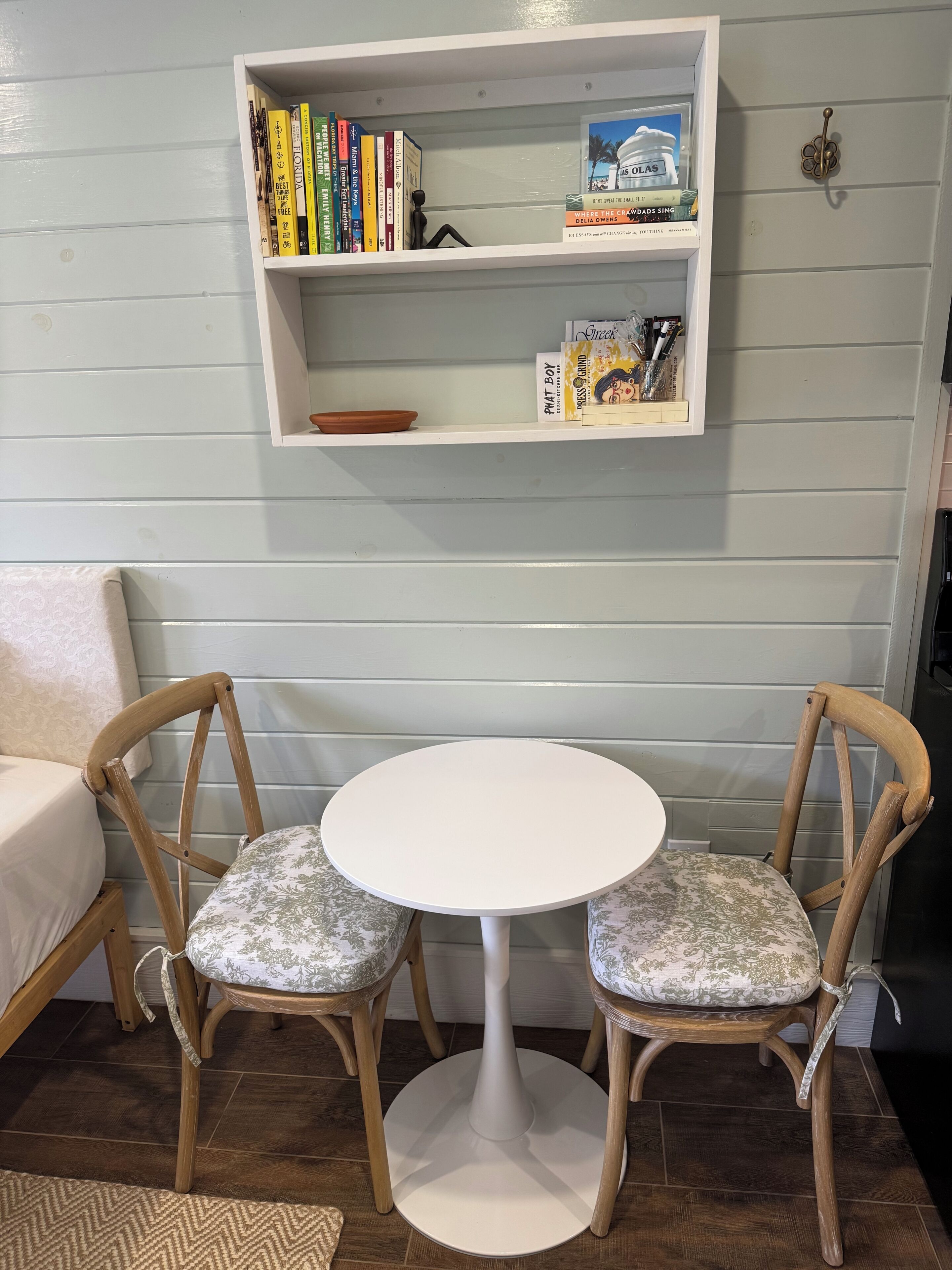 A room with a white round table, two wooden chairs with floral cushions, a bookshelf with books and decorative items, and a light-colored wall.