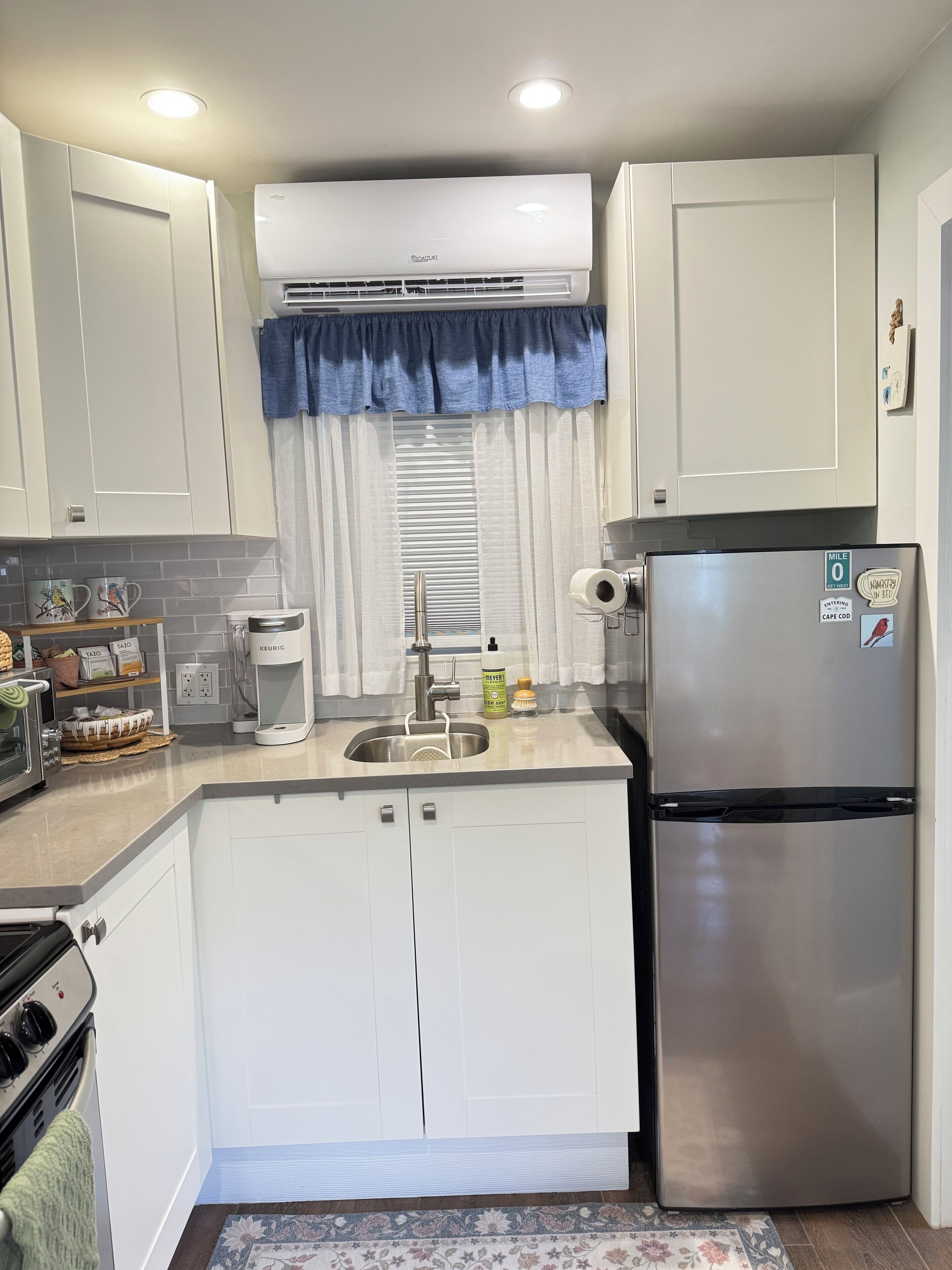 Cocina compacta con armarios blancos, refrigerador de acero inoxidable, aire acondicionado integrado, tetera y cafetera.