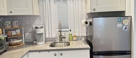 Cocina compacta con armarios blancos, refrigerador de acero inoxidable, aire acondicionado integrado, tetera y cafetera.