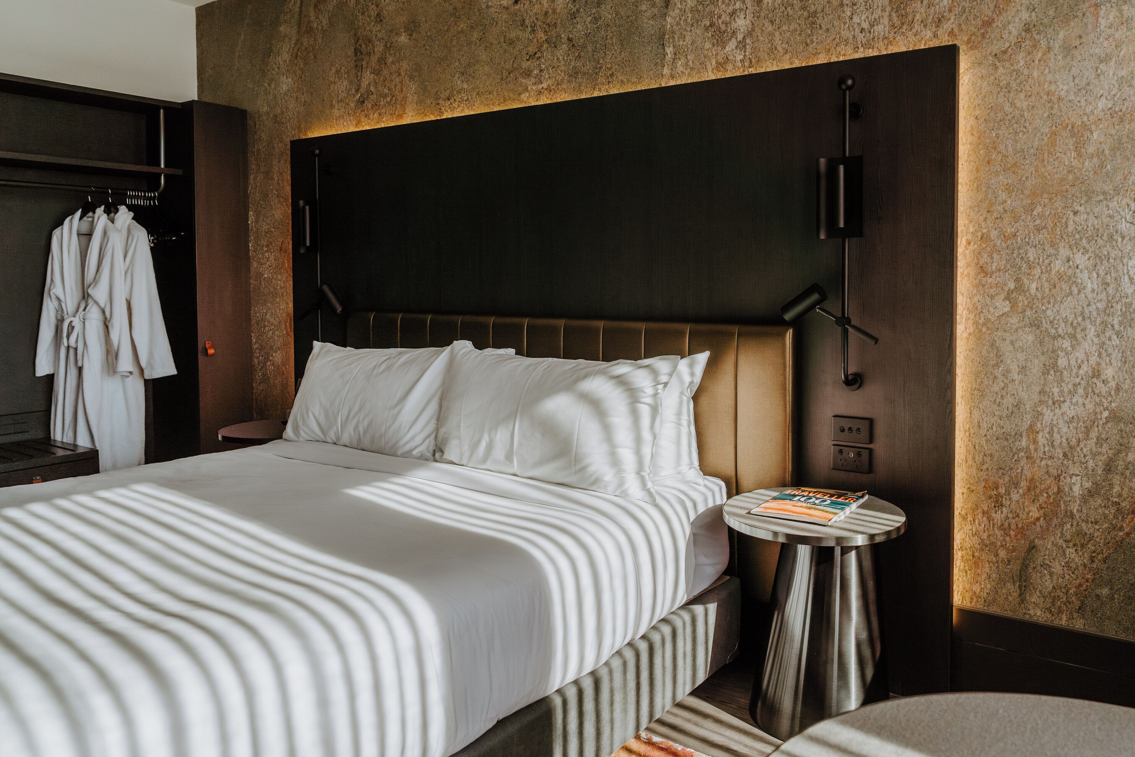A hotel room with a large bed, a bedside table with a magazine, and a wardrobe with hanging robes.