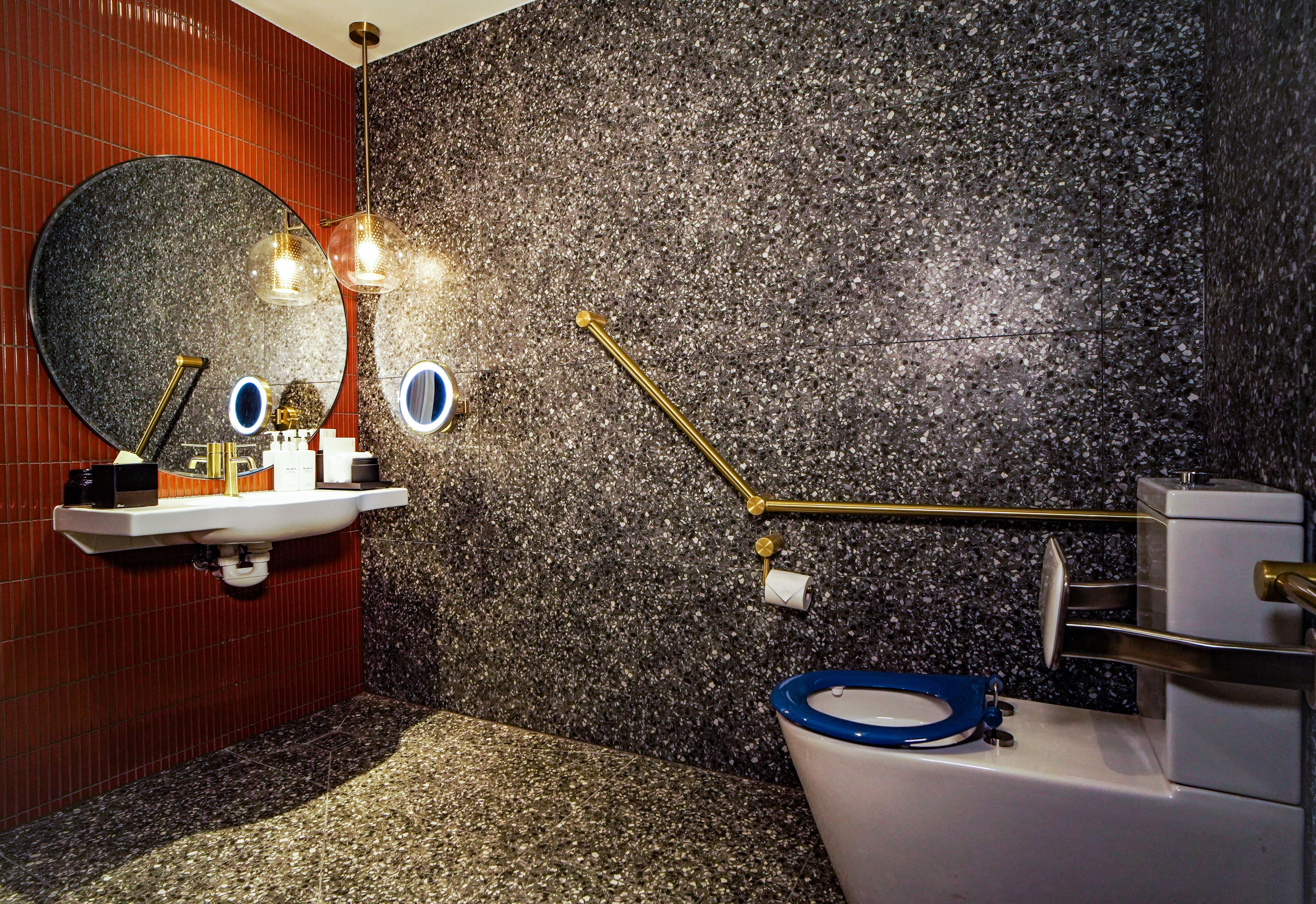 A bathroom with a granite countertop sink, a large round mirror, and a toilet with a blue toilet paper holder.